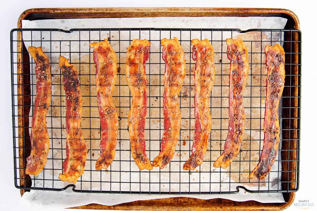 Seven strips of crispy bacon are laid out evenly on a metal rack over a baking sheet lined with parchment paper. The bacon appears well-cooked and is spaced apart.