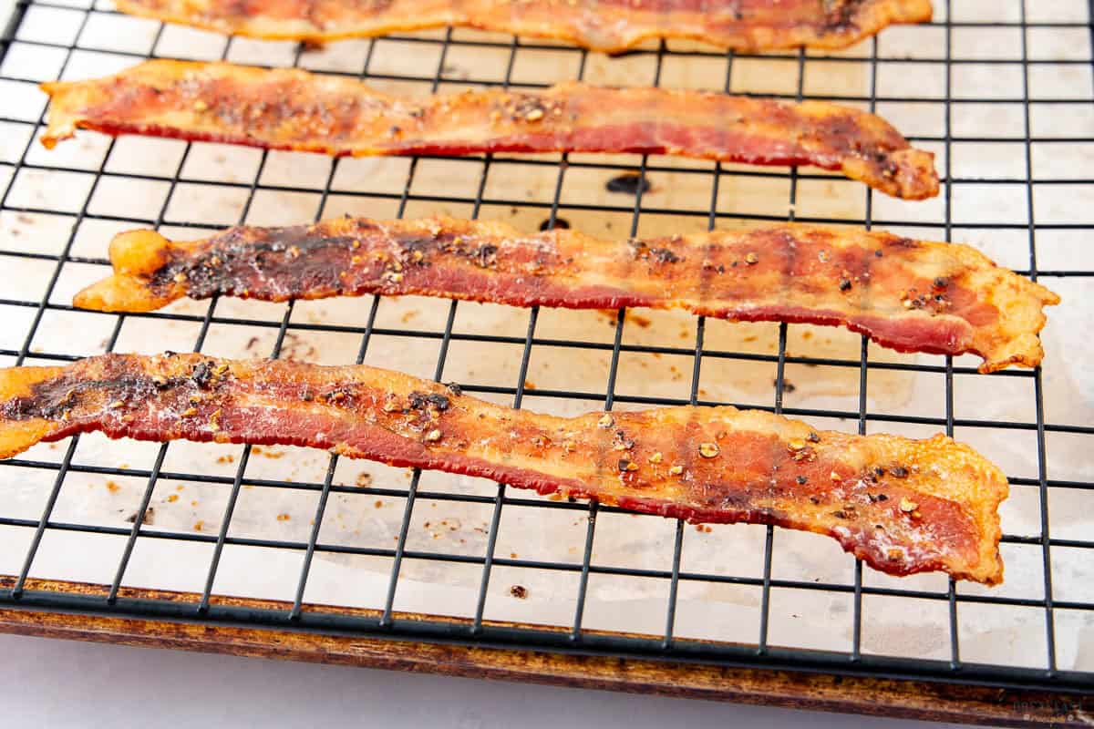 Crispy bacon strips cooling on a wire rack over a baking sheet lined with parchment paper. The bacon is evenly cooked, with a slightly charred appearance and pepper seasoning visible on the surface.