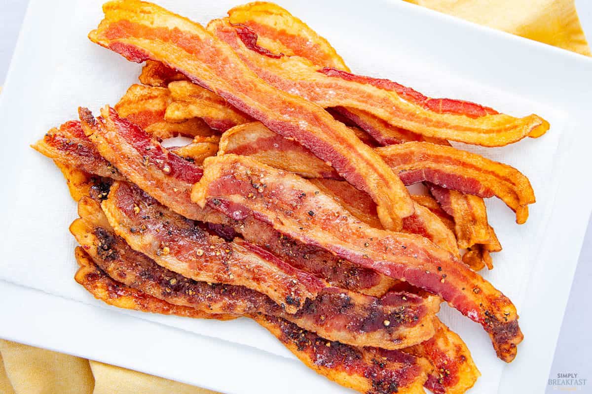 A pile of crispy cooked bacon strips on a white rectangular plate with a paper towel underneath, set on a light yellow cloth. The bacon has a golden brown color with some areas showing seasoning.