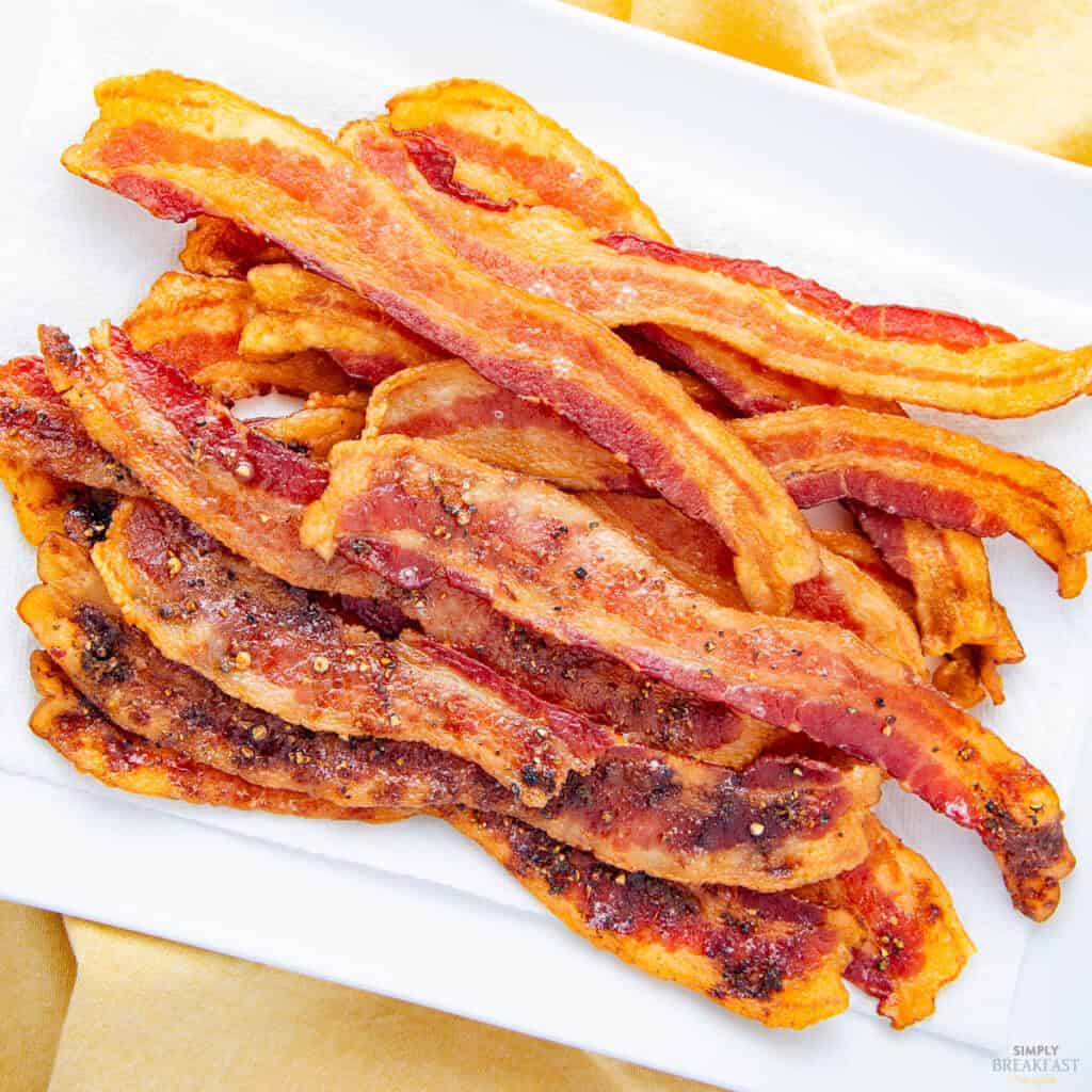 A plate of crispy cooked bacon strips is arranged on a white rectangular dish with a yellow napkin underneath. The bacon is golden brown with a slightly charred texture, suggesting it's well-cooked and seasoned.