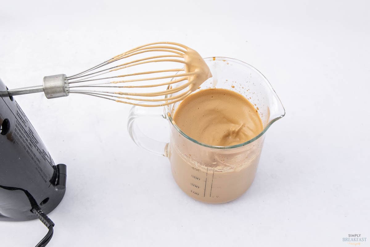 A whisk attachment of a hand mixer holds frothy, light brown whipped coffee over a glass measuring cup filled with the same coffee mixture. The mixer is on the left, and the scene is set against a white background.