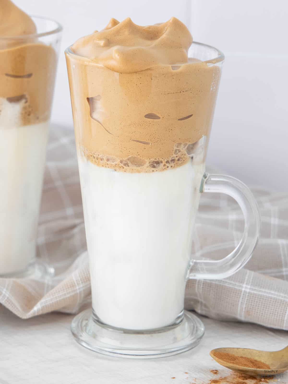 A tall glass of dalgona coffee with creamy, frothy coffee foam on top, over a layer of milk. The glass is placed on a white surface near a light-colored cloth and a small wooden spoon with coffee powder.