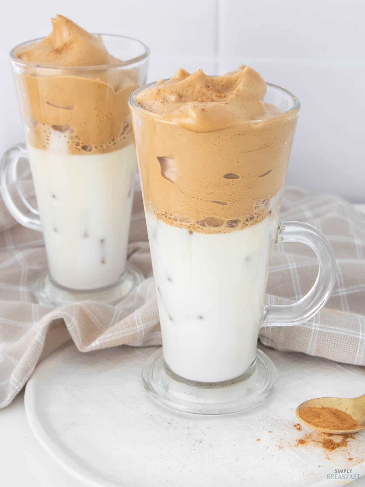 Two tall glasses of dalgona coffee with frothy coffee whip on top and milk below, placed on a white surface. A small amount of cinnamon is sprinkled next to the glasses. A beige cloth lies in the background.