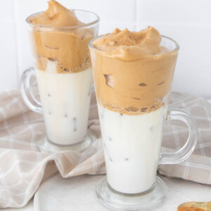 Two clear glass mugs filled with iced milk and topped with whipped coffee foam sit on a checkered beige cloth. The frothy coffee creates a layered look, contrasting with the white milk below.