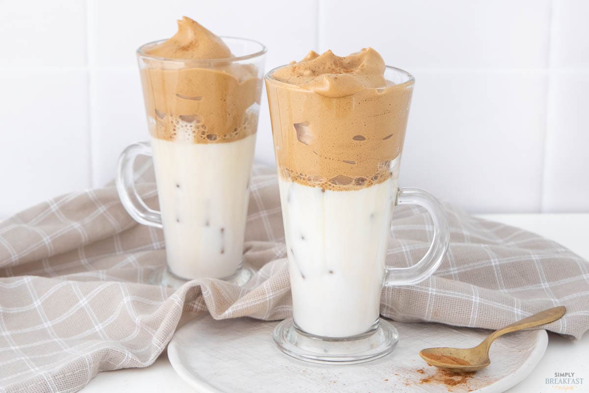 Two glass mugs filled with iced milk and topped with whipped coffee foam, known as Dalgona coffee, are placed on a light-colored napkin. A spoon with coffee powder residue rests nearby. The background is a plain white tile wall.