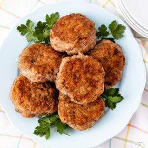 A plate with six golden-brown sausage patties garnished with fresh parsley. The plate is placed on a checkered tablecloth with stacked white plates in the background.