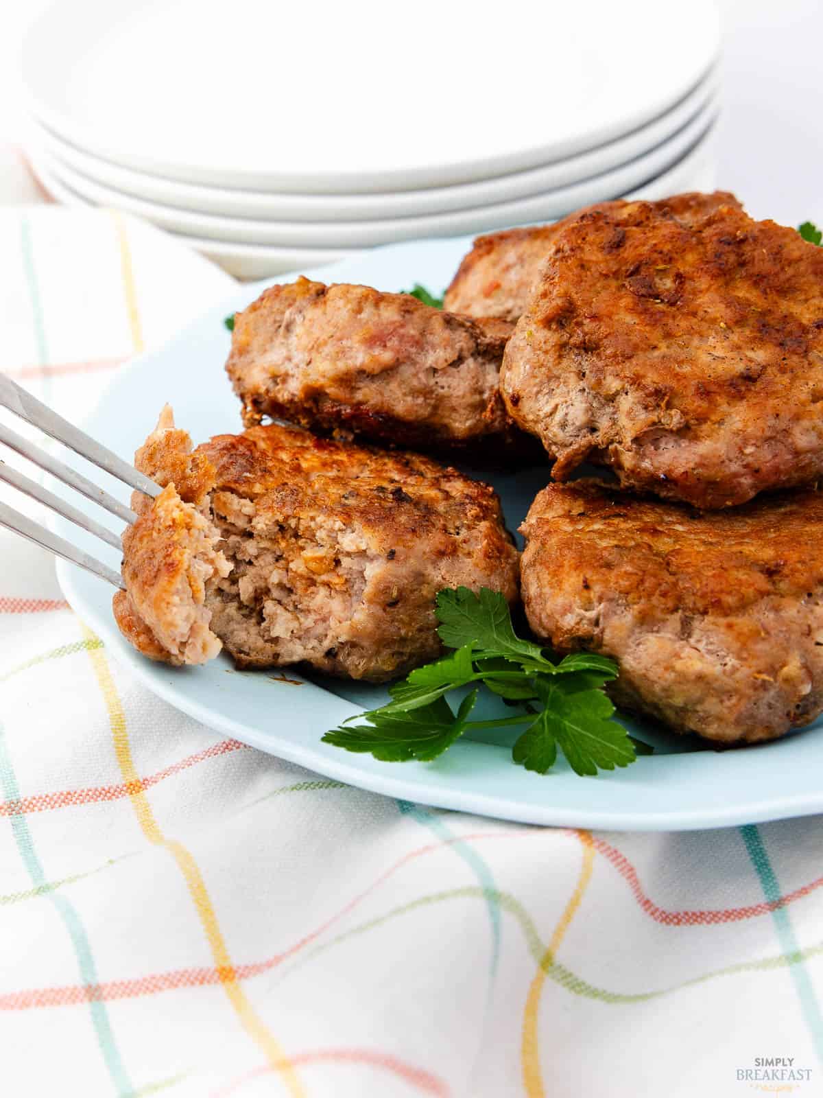 Four browned sausage patties on a light blue plate with parsley garnish. A fork holds one patty with a bite missing. Plates and a colorful striped cloth are in the background.