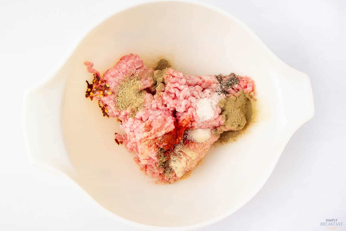 Ground meat seasoned with various spices, including pepper, red pepper flakes, and herbs, is placed in a white mixing bowl. The ingredients are ready to be mixed, set against a plain white background.