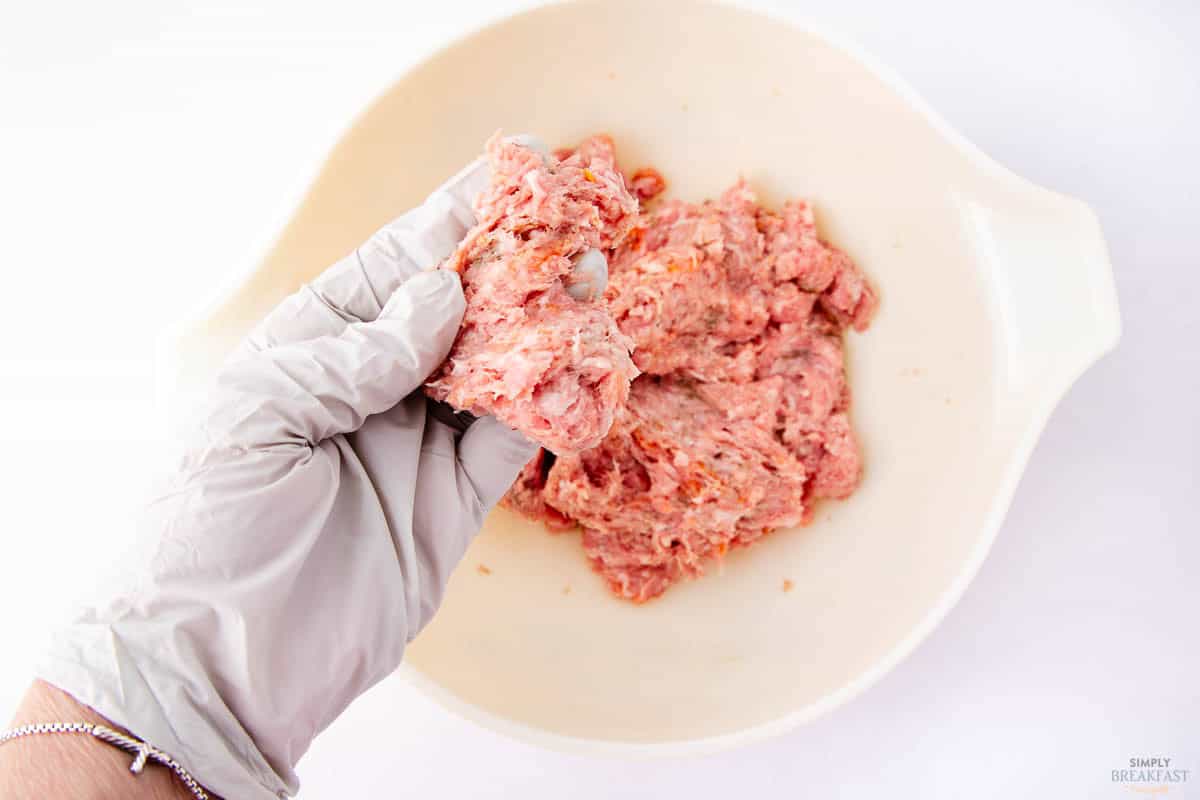 A hand wearing a white glove holds a piece of light pink ground meat over a white bowl filled with a larger quantity of the same meat. The scene is set against a plain white background.