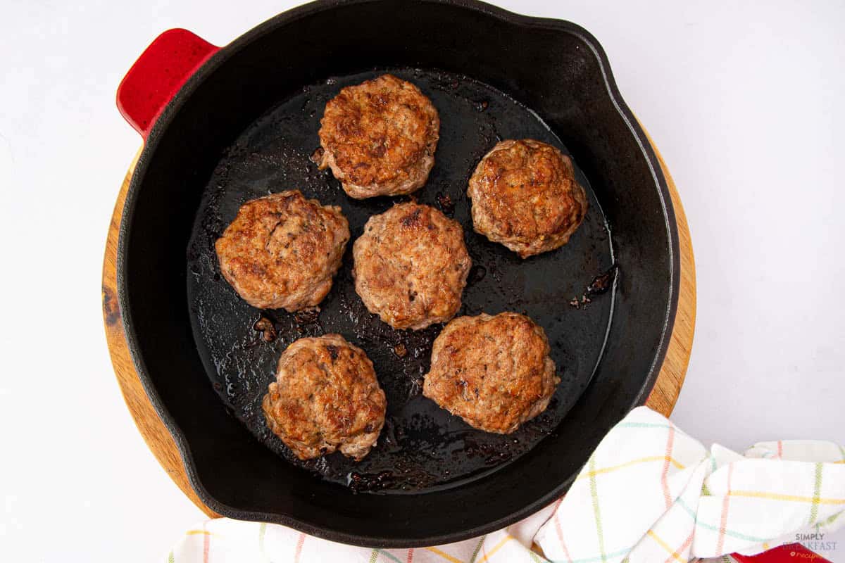 A cast iron pan containing six cooked, browned sausage patties on a wooden trivet. A white and pastel checkered cloth is partially visible in the lower right corner. The background is white.