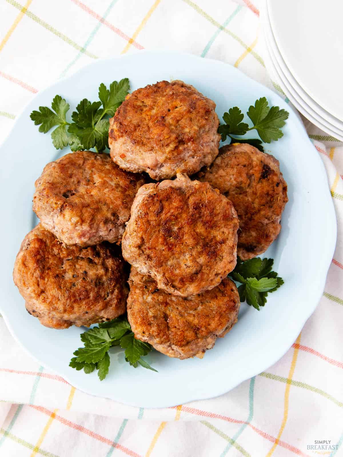 A light blue plate with six grilled sausage patties garnished with fresh parsley on top of a white tablecloth with a colorful check pattern. A stack of white plates is visible in the background.