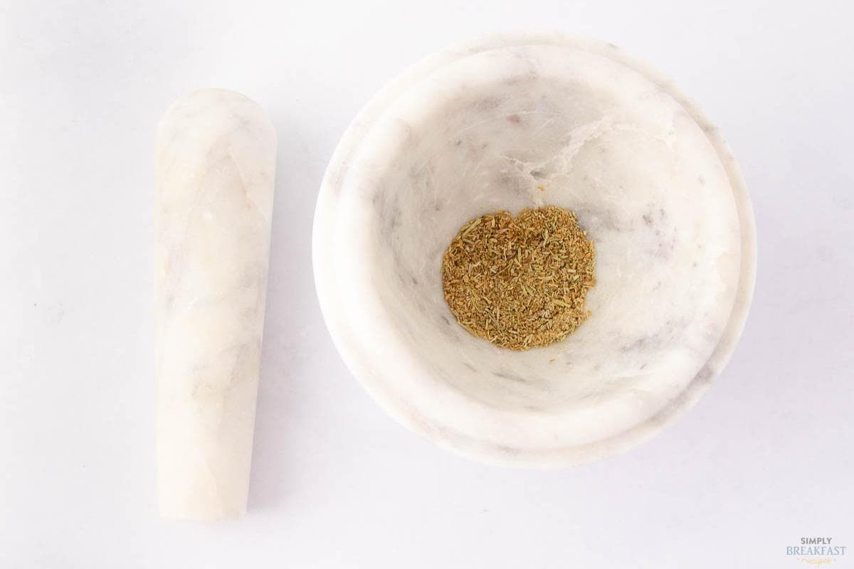 A marble mortar filled with crushed spices sits on a white surface, the perfect start for a savory pork sausage recipe, with a matching pestle resting to the left.