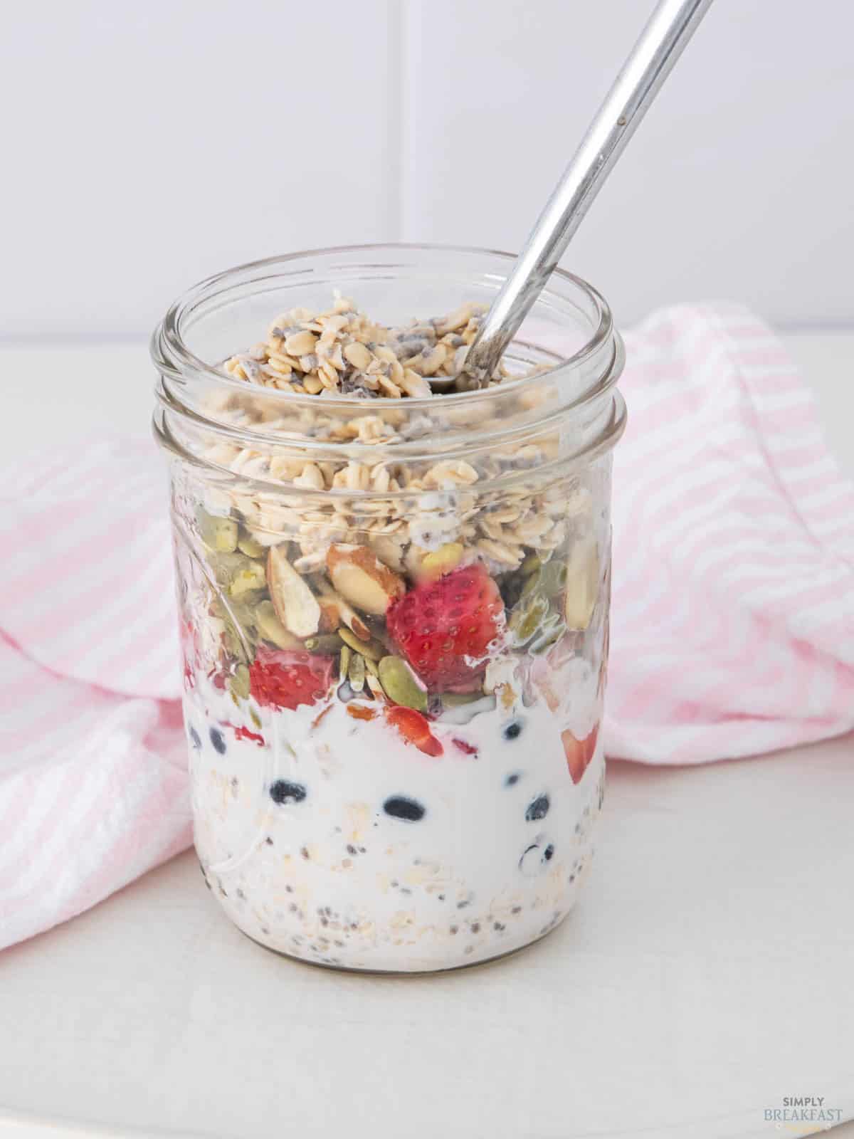 A glass jar filled with overnight oats, topped with sliced strawberries, blueberries, seeds, and nuts. A spoon is inside the jar, and a pink striped cloth is in the background.