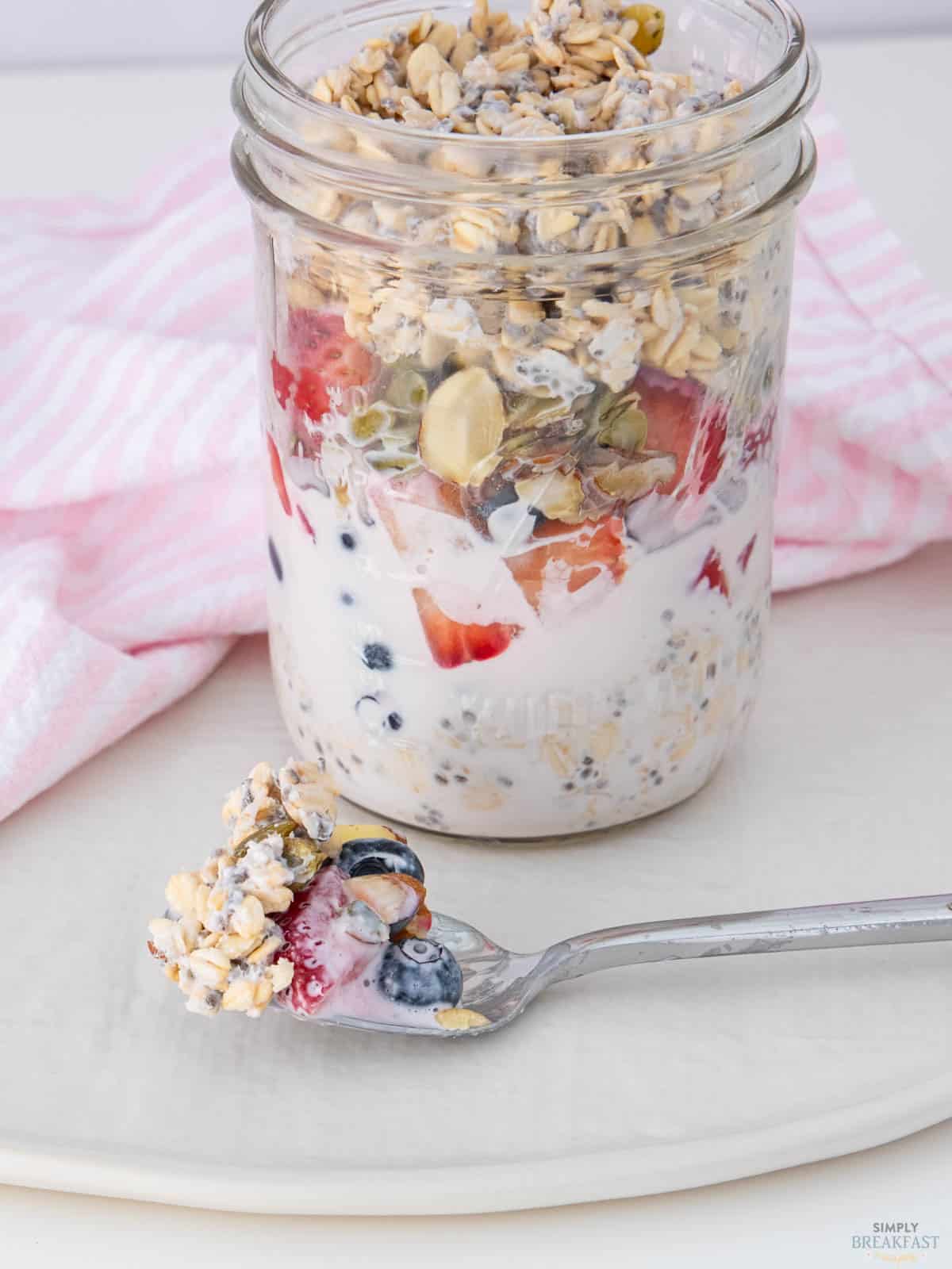 A mason jar filled with layered oats, yogurt, sliced strawberries, blueberries, and granola. A spoon with more of the mixture rests on a white plate beside the jar. A pink and white striped cloth is in the background.