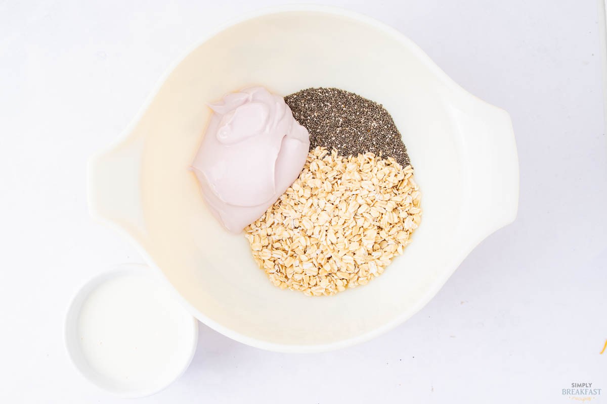 A white bowl contains a portion of yogurt, chia seeds, and oats. A small white cup with milk is next to the bowl. The ingredients are arranged separately, ready for mixing.