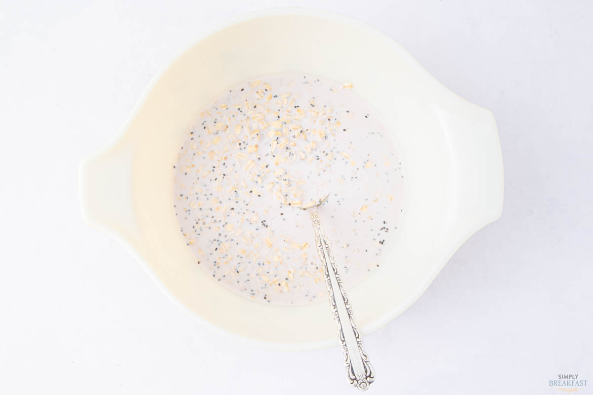 A white bowl with handles contains a mixture of oats, chia seeds, and milk. A silver spoon rests in the bowl. The background is a light surface, enhancing the simplicity and freshness of the image.