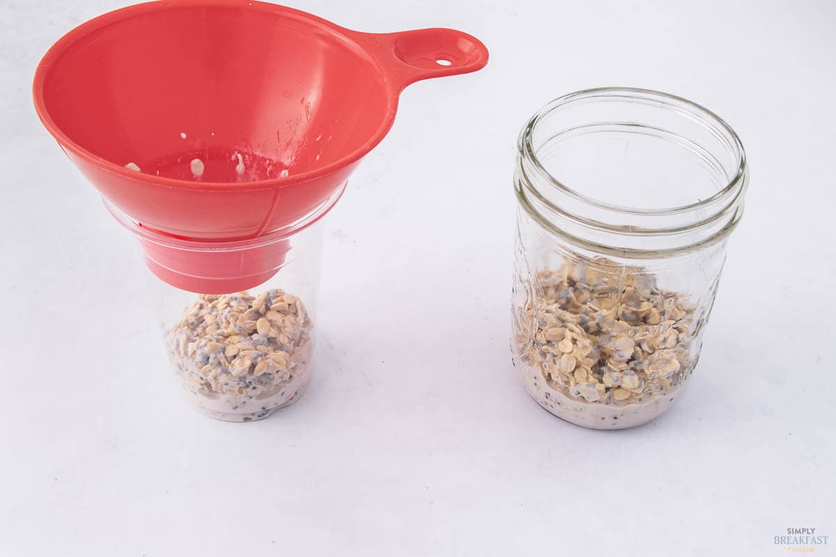 A red funnel is placed over a glass jar partially filled with oatmeal. Another jar beside it contains a similar oatmeal mixture. Both jars are on a white surface.