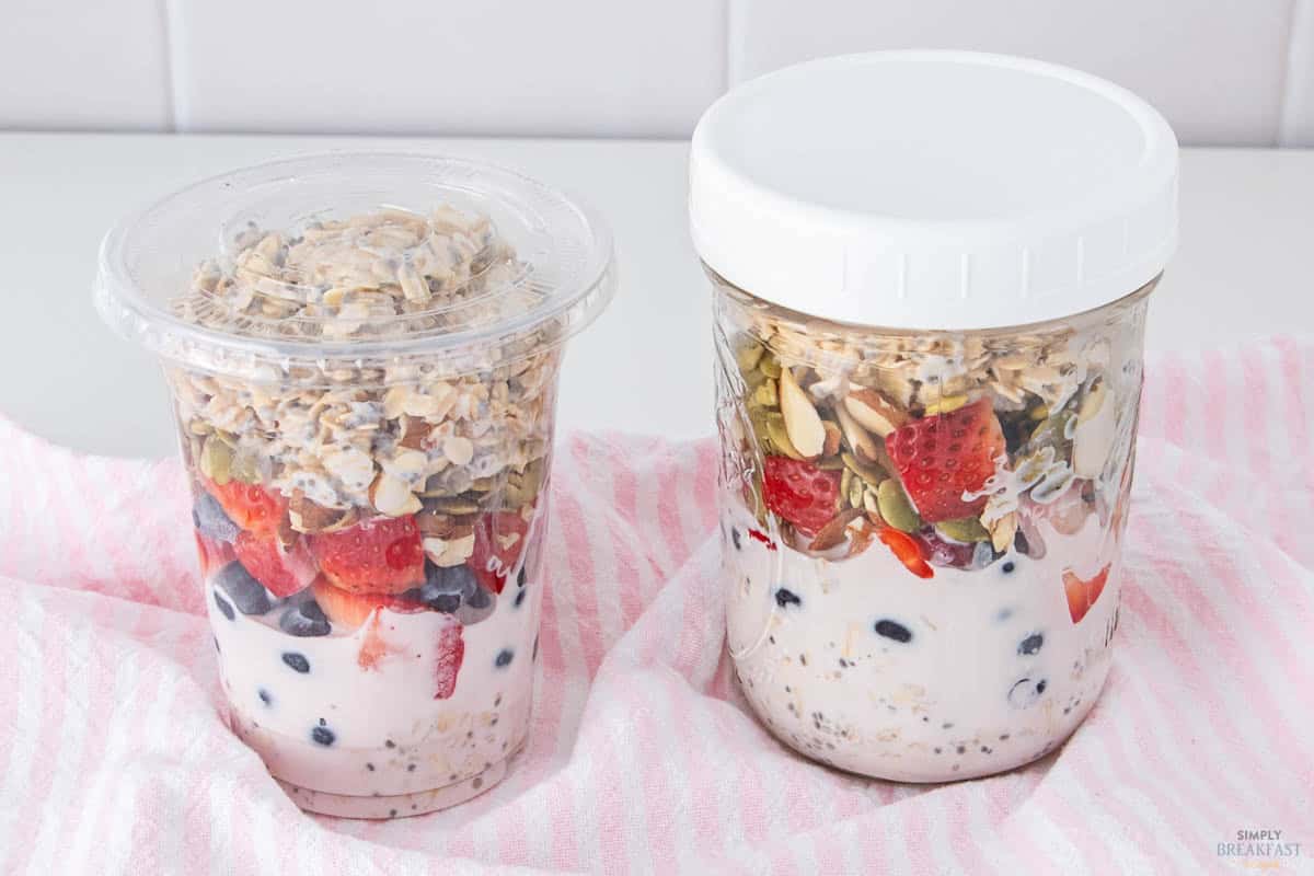 Two jars of overnight oats sit on a pink striped cloth. Both jars contain layers of yogurt, blueberries, sliced strawberries, oats, and seeds. The jar on the left has a clear lid, while the one on the right has a white lid.