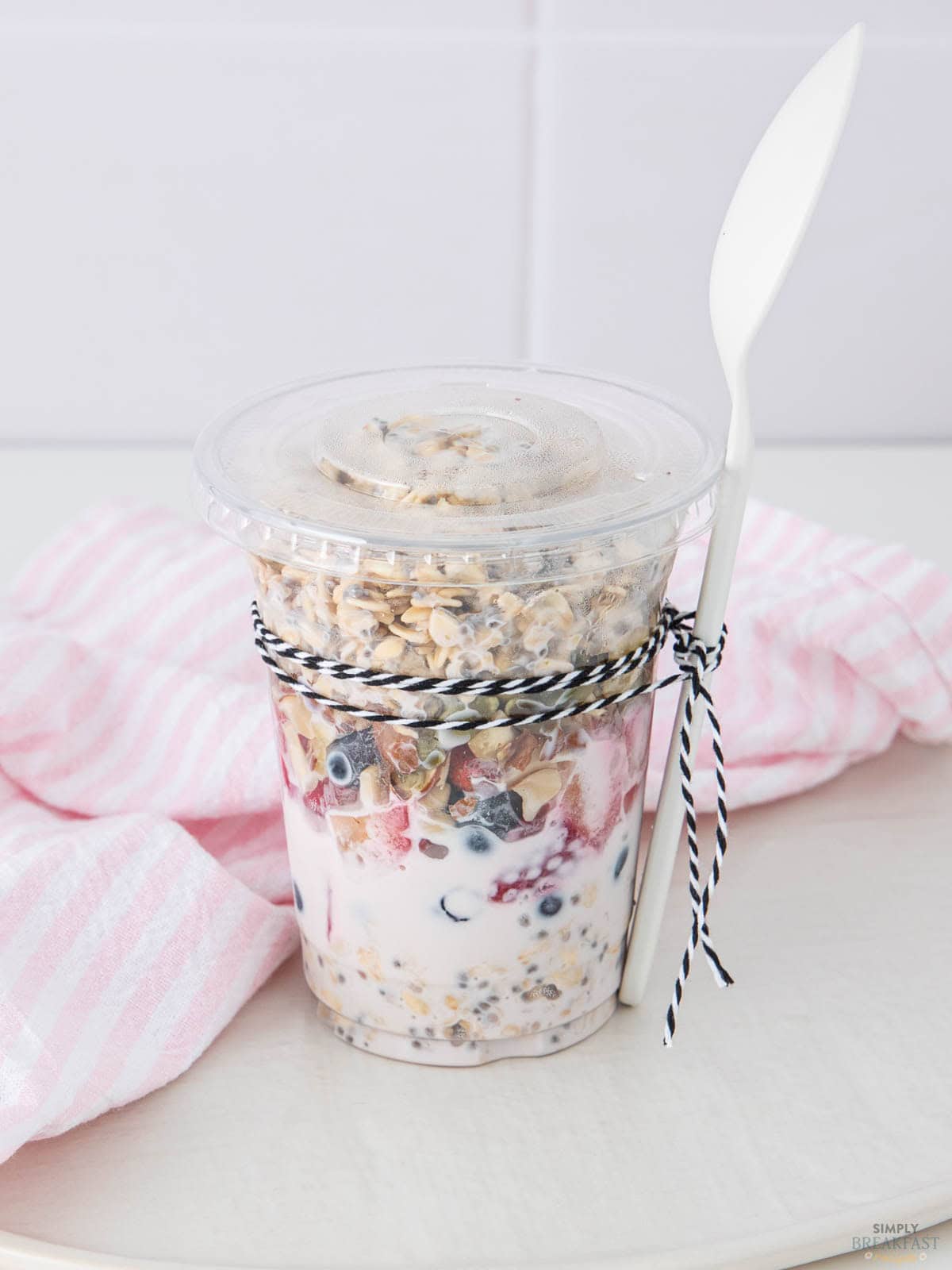 A parfait in a clear plastic cup filled with layered granola, yogurt, and blueberries. It's topped with a granola mix. A black and white string is tied around the cup, securing a white plastic spoon. A pink and white striped cloth is in the background.