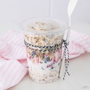 A plastic cup filled with a yogurt parfait, featuring layers of yogurt, granola, and berries. The cup is tied with a black and white string and comes with a white spoon. A pink striped cloth is in the background.