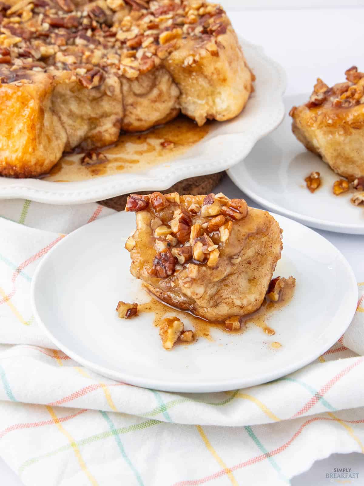 A piece of sticky bun topped with chopped nuts is placed on a small white plate in the foreground. Behind it, more sticky buns sit on a larger platter. A colorful striped cloth is partially visible underneath.