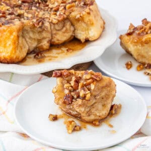 A piece of sticky bun topped with glazed pecans on a white plate. The background shows additional sticky buns on a serving platter. A checkered cloth is partially visible underneath the plates.