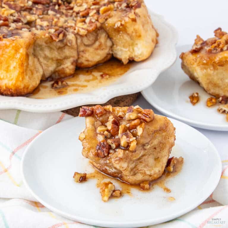 A piece of sticky bun topped with glazed pecans on a white plate. The background shows additional sticky buns on a serving platter. A checkered cloth is partially visible underneath the plates.