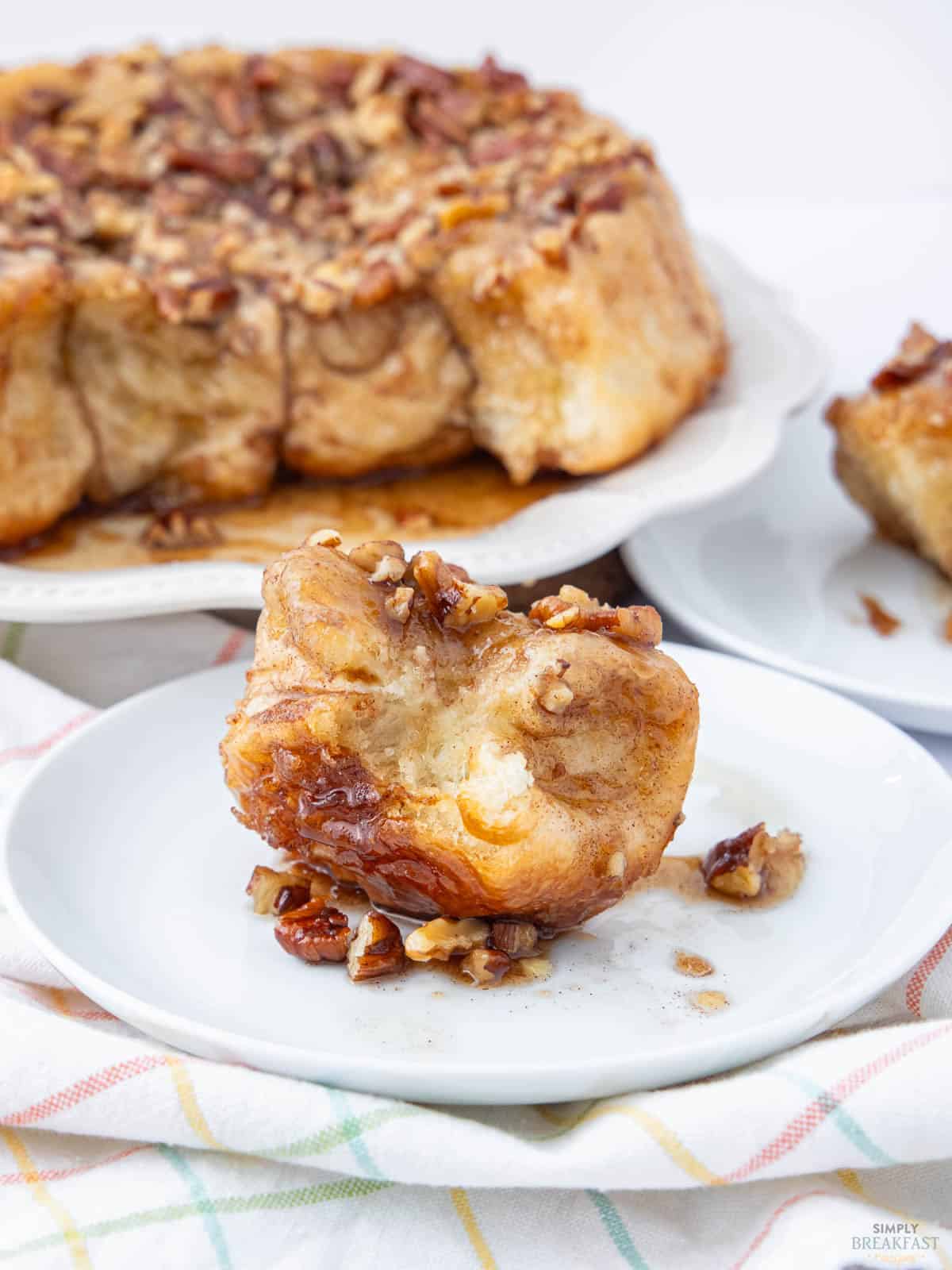 A sticky bun topped with chopped pecans sits on a white plate. In the background, more sticky buns can be seen on a platter. The table is covered with a pastel plaid cloth.