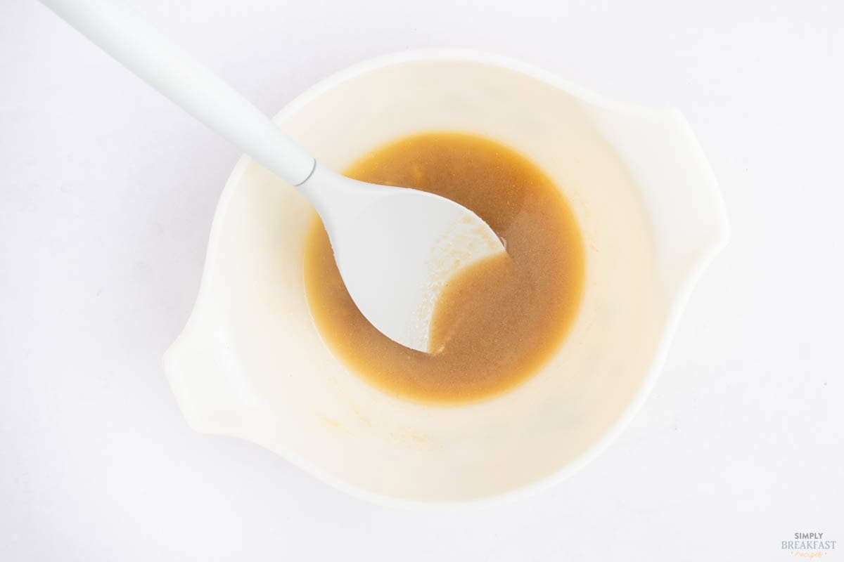 A white bowl containing brown liquid with a white spatula resting inside. The bowl is against a plain white background.