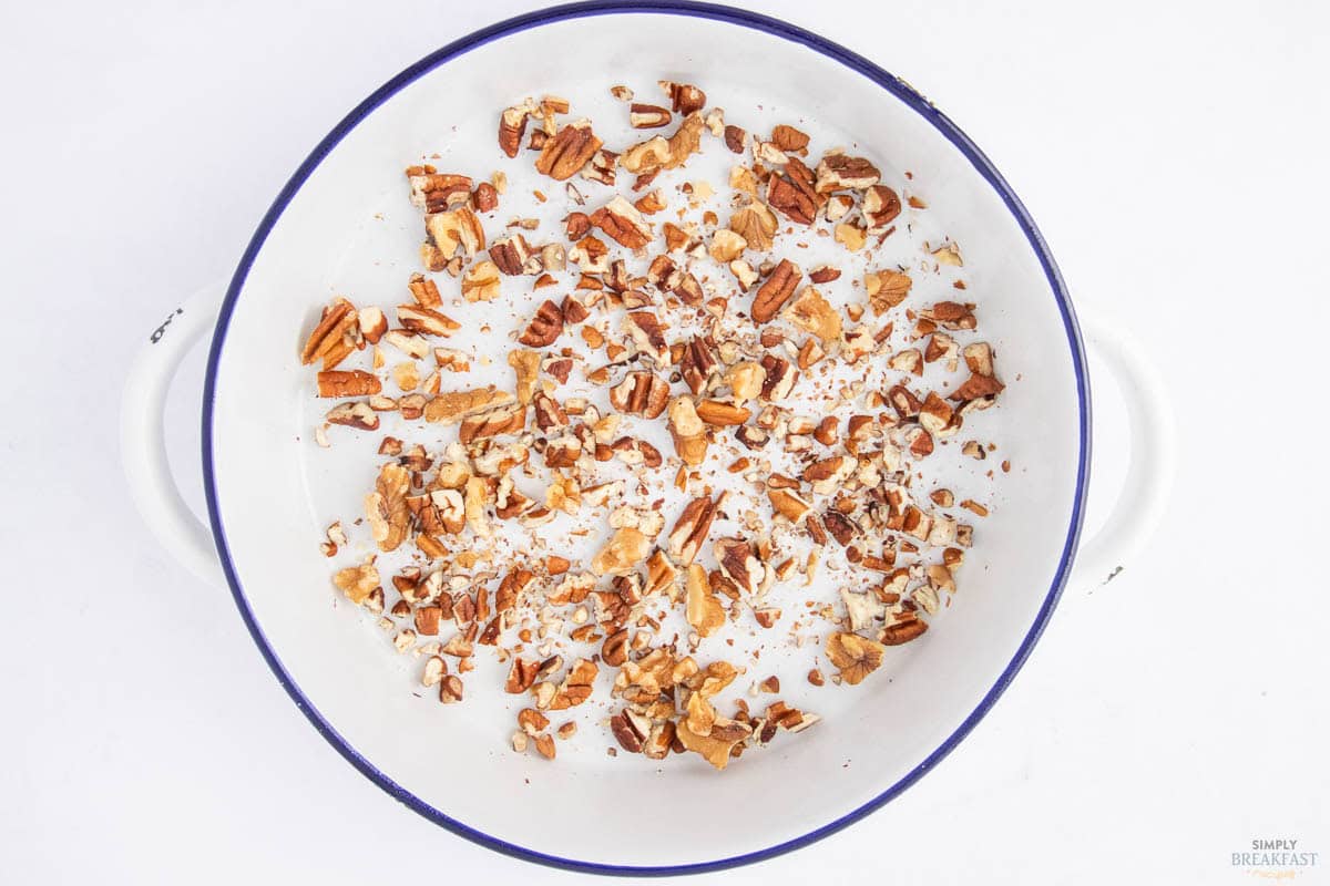 A round white dish with a dark rim, filled with chopped pecans spread evenly across the bottom. The background is a plain white surface.