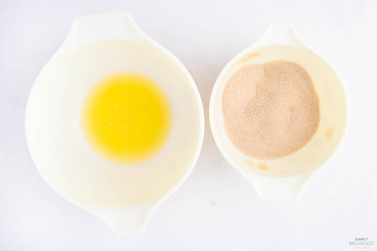 Two white bowls on a white surface. The left bowl contains melted butter, and the right bowl contains cinnamon sugar.