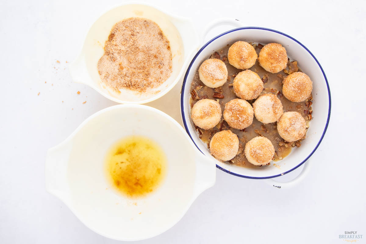 Baking dish with round biscuits covered in cinnamon sugar on a bed of pecans; next to it are two bowls: one with melted butter and the other with cinnamon sugar mix.