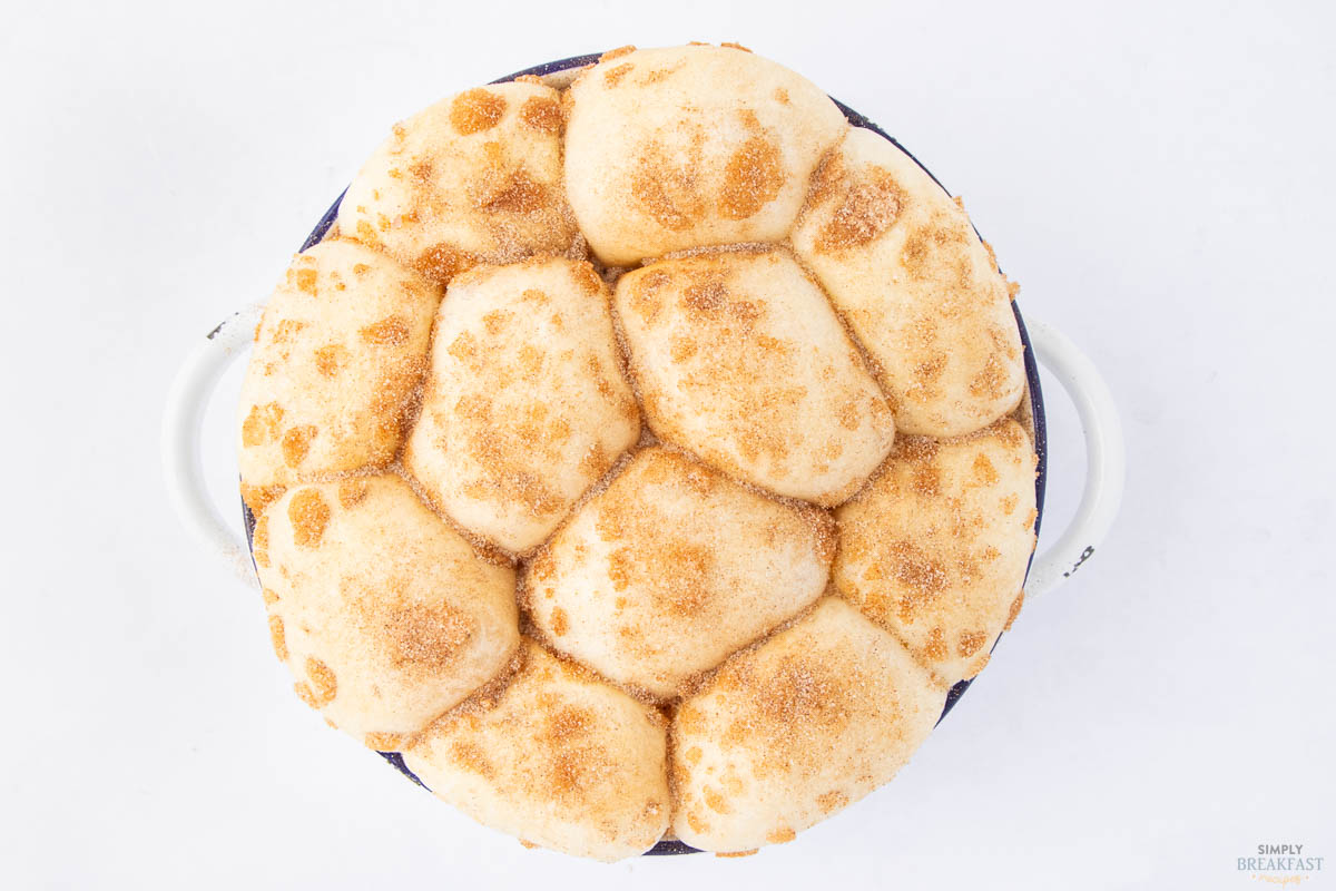 A top view of a pull-apart bread loaf covered with cinnamon sugar, in a round baking dish with handles. The bread is golden brown and arranged in a clustered pattern, on a white background.