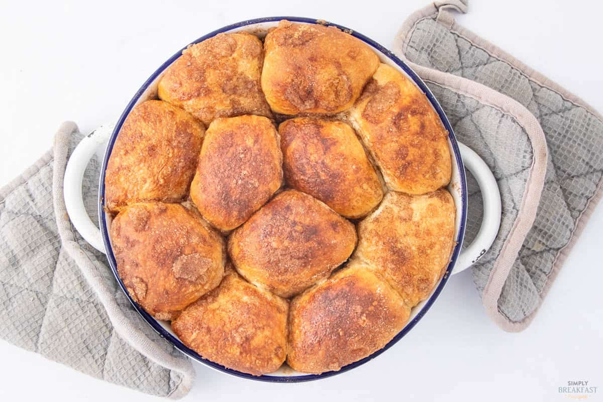 A round pan holds freshly baked cinnamon rolls, with a golden-brown crust. The pan is set atop two quilted grey oven mitts, one on each side, on a white surface.