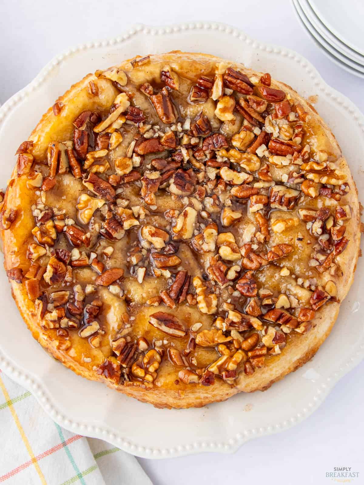 A round, caramel-glazed sticky bun topped generously with chopped pecans, walnuts, and almonds sits on a decorative white plate. A plaid napkin is partially visible beside the plate on a white surface.