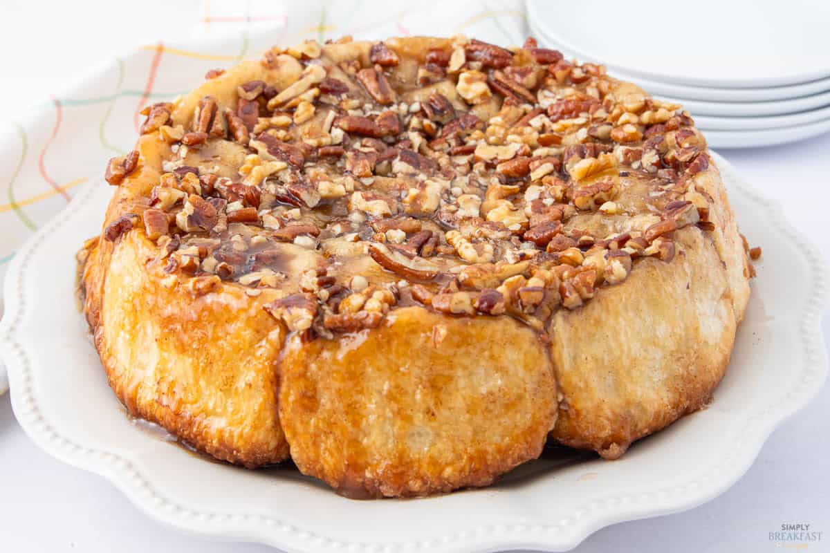 A caramel pecan sticky bun cake with a glossy glaze, topped with chopped pecans. The cake is on a white, scalloped plate. In the background, there is a colorful striped cloth and stacked white plates.