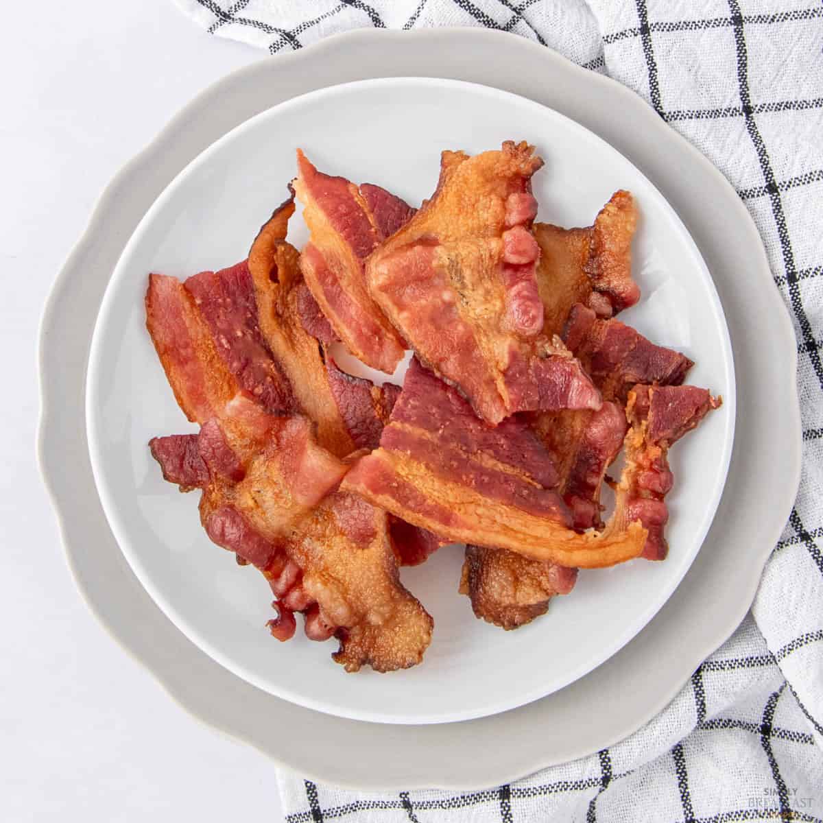 A white plate filled with several crispy, cooked bacon strips sits on a gray-edged plate, placed on a white and black checkered cloth.