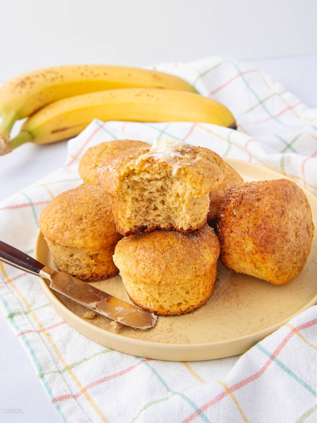 Une assiette de muffins à la cannelle, dont un avec une bouchée prise et du beurre qui fond sur le dessus, est posée sur un tissu écossais à côté d'un couteau à beurre et de bananes mûres en arrière-plan.