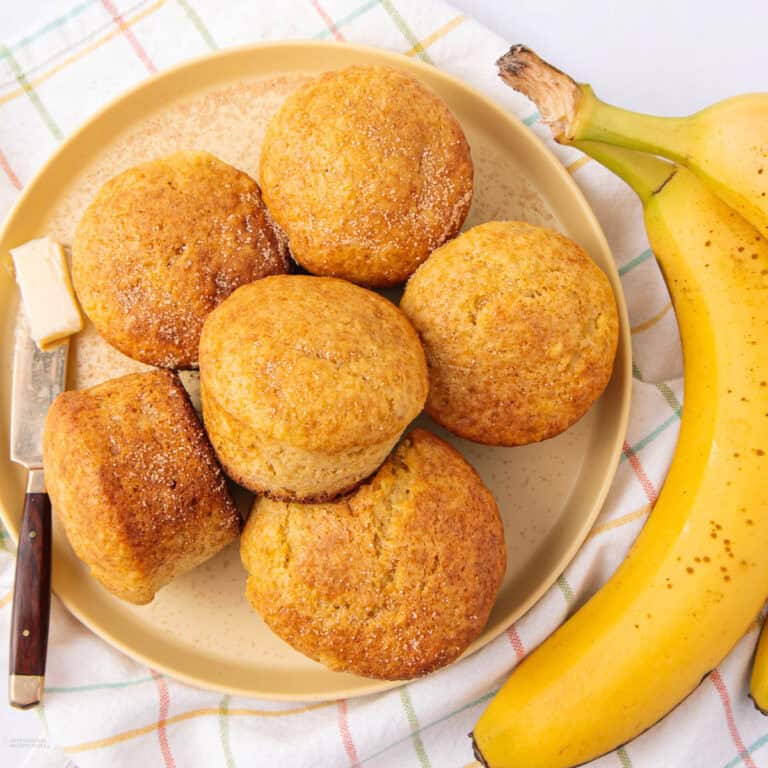 A plate with six golden brown muffins, a butter knife with a pat of butter, and two ripe bananas, all set on a plaid cloth.
