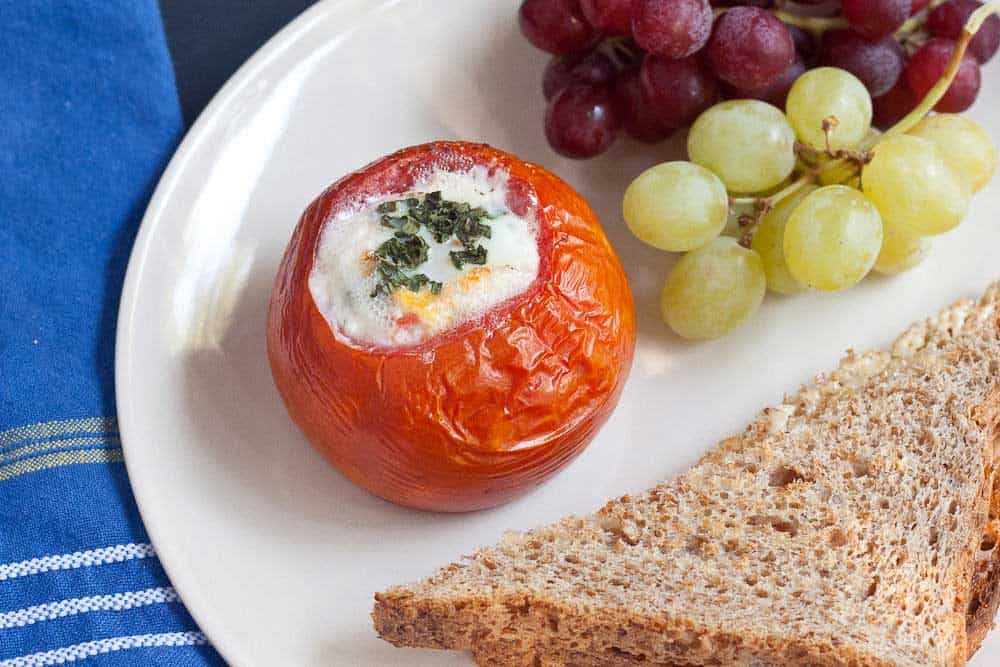 A stuffed baked tomato with baked eggs and herbs, served with red and green grapes and a slice of whole wheat bread on a white plate.