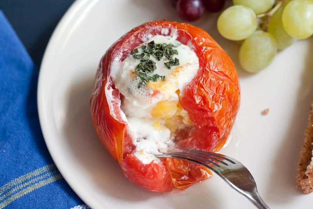 A baked tomato filled with savory baked eggs and garnished with herbs on a white plate, served alongside green grapes and a slice of bread.