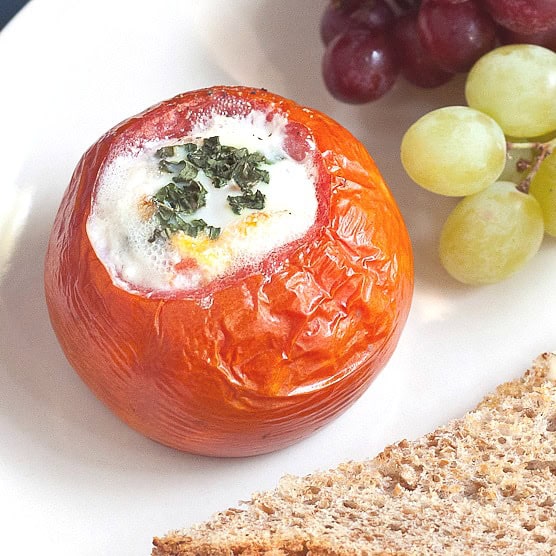 A baked tomato stuffed with egg and herbs sits on a plate, served with green and red grapes and a slice of brown bread—a flavorful choice for breakfast recipes.
