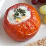 A baked tomato filled with baked eggs and topped with chopped herbs, served on a plate with grapes and a slice of bread.
