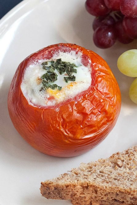 A baked tomato filled with baked eggs and topped with chopped herbs, served on a plate with grapes and a slice of bread.