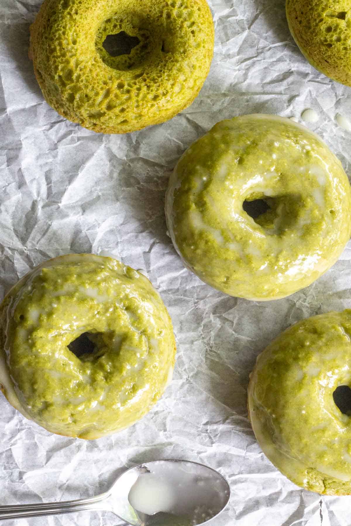 Three baked matcha donuts glazed in green, along with one unglazed doughnut, rest on crumpled parchment paper beside a metal spoon holding white glaze.