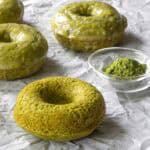 Three baked matcha donuts with a glossy matcha glaze and one plain baked matcha donut rest on crumpled parchment paper next to a small glass bowl of matcha powder.
