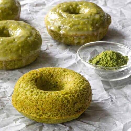 Three baked matcha donuts with a glossy matcha glaze and one plain baked matcha donut rest on crumpled parchment paper next to a small glass bowl of matcha powder.