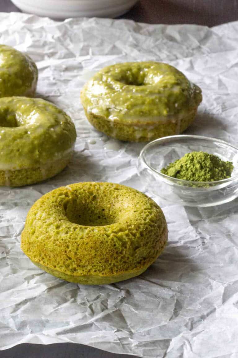 Three baked matcha donuts with a glossy matcha glaze and one plain baked matcha donut rest on crumpled parchment paper next to a small glass bowl of matcha powder.