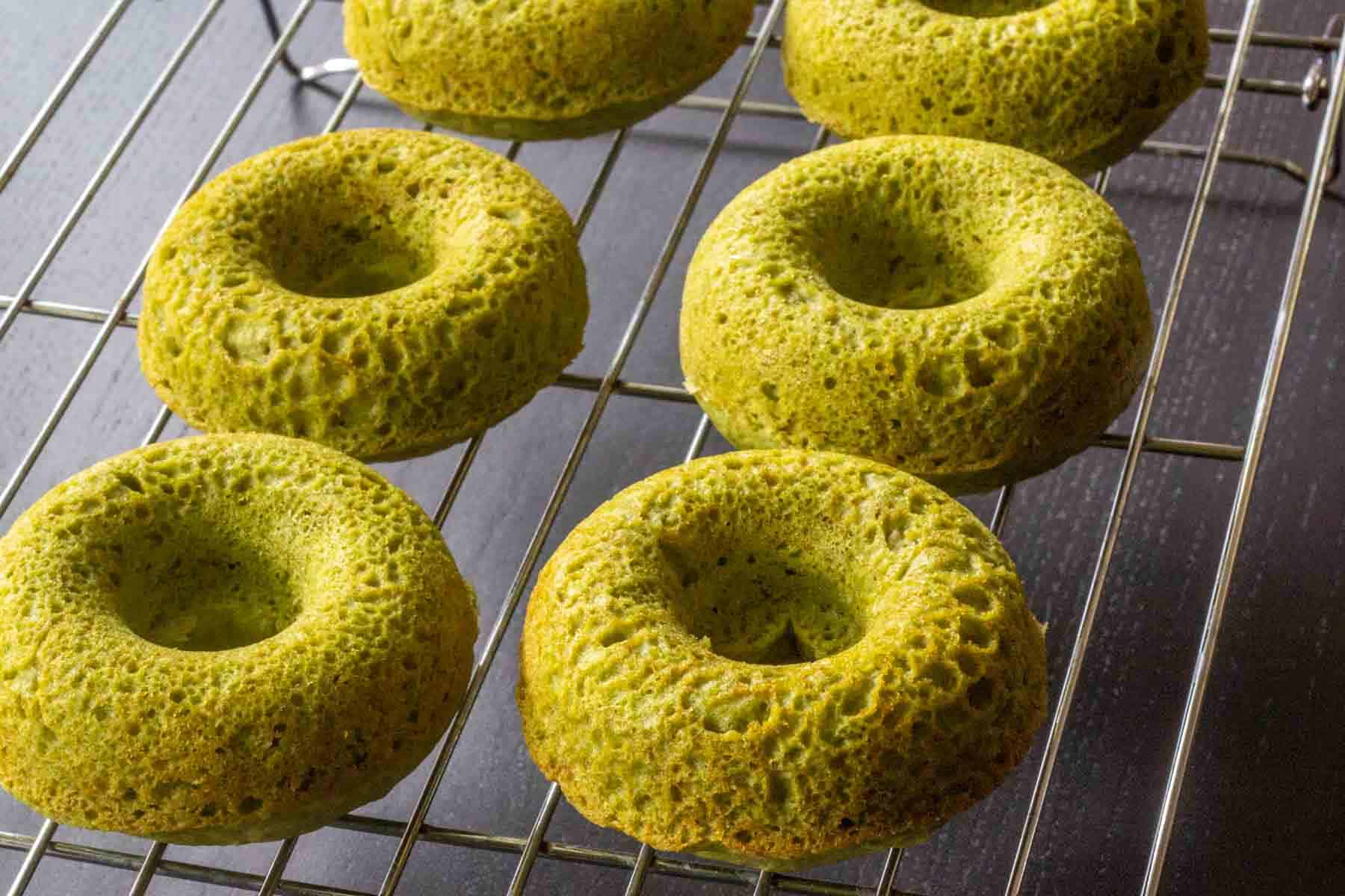 Six baked matcha donuts cool on a metal wire rack atop a dark surface.