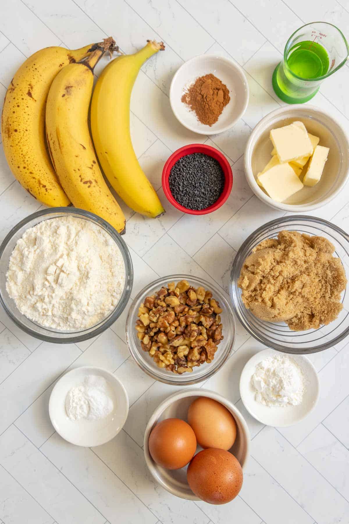 Overhead view of banana nut bread ingredients—bananas, eggs, flour, brown sugar, walnuts, butter, poppy seeds, baking powder, baking soda, cinnamon—and a small glass of green liquid on a white surface.